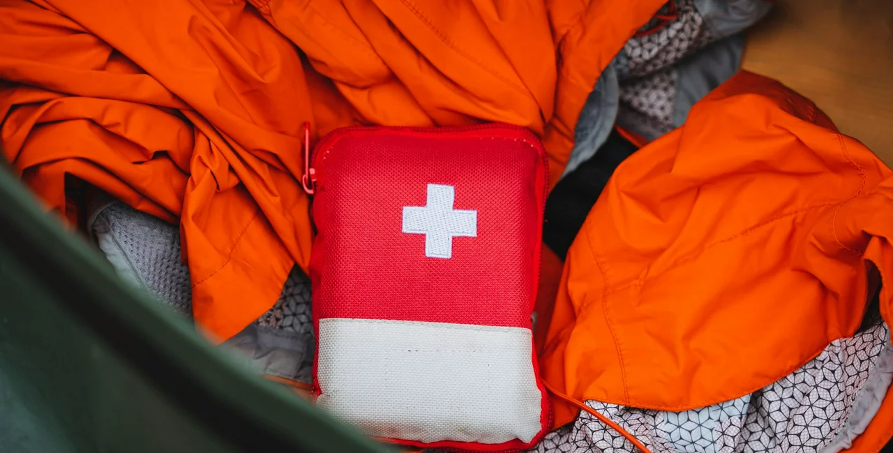 A red first aid kit with a white cross is shown from above, surrounded by bright orange safety gear. It contains essential emergency supplies for outdoor